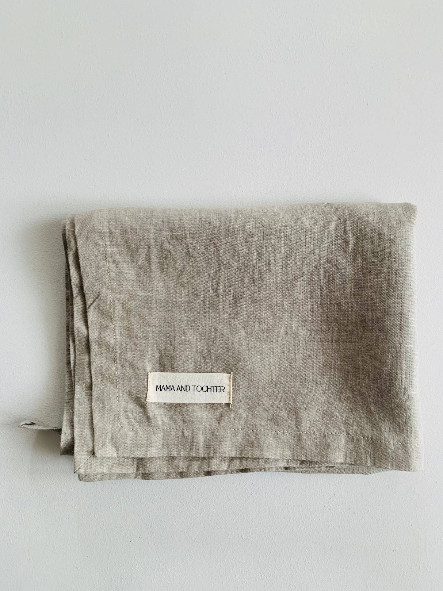 A folded taupe-colored pure French linen tea towel with a visible brand label on the bottom right corner